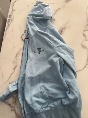 Ljusblå hoodie från Tommy Hilfiger - Snygg ljusblå hoodie från Tommy Hilfiger med dragkedja framtill och fickor på sidorna. Märkets logga är broderad på bröstet. Hoodien har en klassisk huva och är tillverkad i mjukt bomullsmaterial. Perfekt för en avslappnad och stilren look.