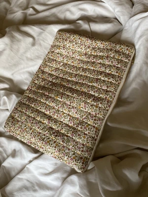 Blommig quiltad datorväska - Säljer en quiltad datorväska med blommigt mönster i beige, rosa, grönt och gult. Väskan har dragkedja längs sidan och är mjukt vadderad för extra skydd. Perfekt för dig som vill ha en söt och unik look till din laptop.