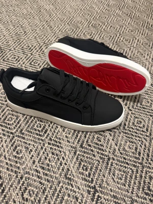 Svarta sneakers med röd sula - Snygga svarta sneakers med vit sula och cool röd undersida. Skorna har en stilren design med svarta snören och diskret logga på hälen. Materialet ser ut att vara en mix av syntet och textil, vilket ger en modern look.