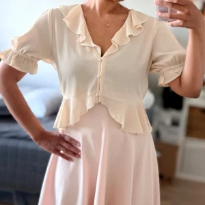 Krämvit corquette blus med volang och knappar - Supersöt krämvit blus med corquette aesthetic från Gina Tricot. Volangdetaljer längs krage och nederkant, korta puffärmar med resår och knappar framtill. Tillverkad i ett lätt, luftigt tyg som ger en romantisk vibe. Perfekt att styla med både kjol och jeans. 