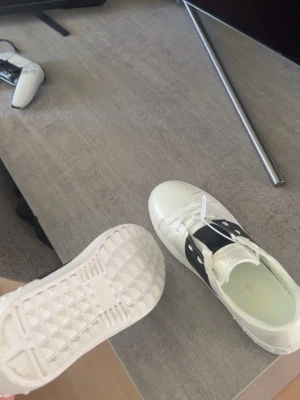 Valentino Garavani vita sneakers - Snygga vita sneakers från Valentino Garavani med svart detalj på sidan och guldfärgad logga på plösen och innersulan. Skorna har klassisk låg profil, vita snören och robust gummisula. Perfekta för dig som vill ha en clean och exklusiv look.