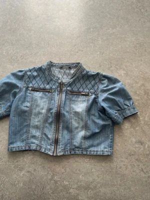 Jeanstopp från Ofelia storlek L - Säljer en jeans topp med dragkedja framtill och två bröstfickor med dragkedja. Toppen har puffiga korta ärmar och ett snyggt quiltat mönster över axlarna. Perfekt för dig som vill ha en unik denimlook.