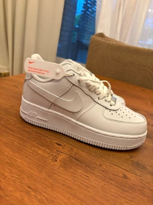 Nike Air Force 1 vita sneakers - Klassiska Nike Air Force 1 sneakers i helvitt läder med vit sula och vita skosnören. Ikonisk swoosh-logga på sidan och perforeringar på tån för extra ventilation. Stilren och tidlös modell som passar till det mesta.
