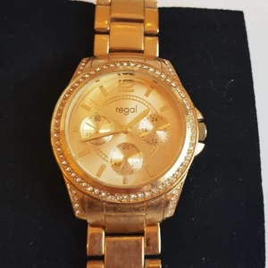 Regal Damklocka - Guldfärgad  - Snygg & Elegant analogklocka från Regal. Damklocka i mycket fint skick, är nästan aldrig använd. Legat fint förvarad och i princip fortfarande som ny. Klockan är Guldfärgad med vackra dekorativa stenar av Swarovski. ONESIZE men är justerbar. Fås i en fin presentask. 🛍️🎁                                                               Nytt batteri ingår.                                                    Fler bilder kan skickas vid intresse.                     Ordinariepris, 449:- 