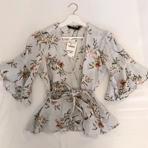 Blus från Zara  - Blus/tunn kofta från ZARA. Oanvänd, prislappen sitter kvar. Volanger i ärmarna och knytband i midjan. Ord. pris 399kr.