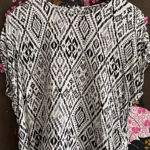 Mönstrad blus/tunika m. omlottrygg - Kortärmad, mönstrad blus/tunika  med veck i omlott på ryggen. Söt virkad kant på rygg och i nederkanten. Uppskattar den till en S, lappen var bortklippt när jag köpte den begagnat. I fint skick!