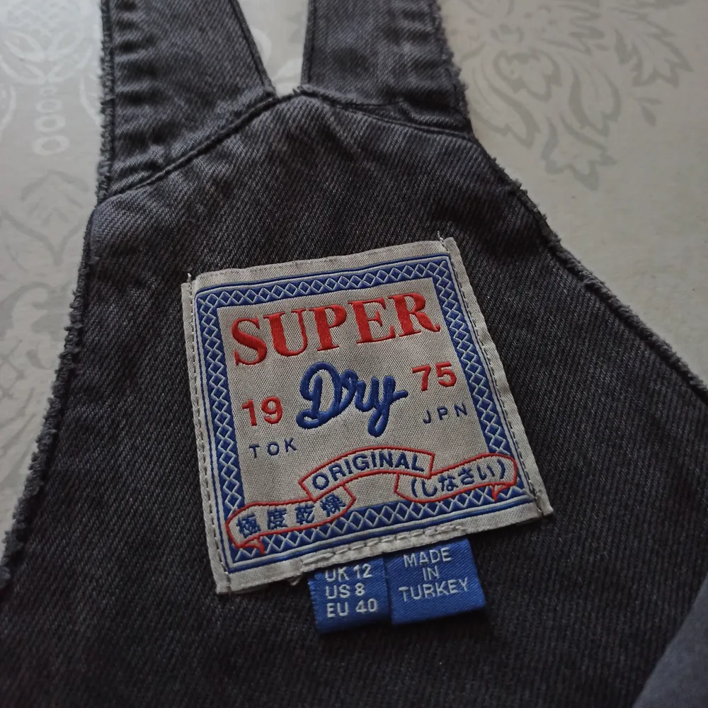 Ett par grå/svarta hängselbyxor i jeansmaterial ifrån Superdry med revor på benen. Byxorna är väl 