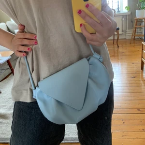 Söt blå väska! - Väskan är inte alls gammal, ungefär 6 månader kanske. Helt oanvänd och i bra skick! Ljusblå färg med skinn-liknande material. Väskan har ett axelband samt knapp och kommer från ”MANGO”