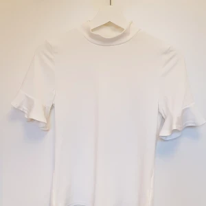 Ribbad top - Vit, ribbad, jättefin T-shirt/topp med små volanger på ärmarna. Nyskick, helt oanvänd och säljes pga att den blev för stor på mig som är mellan strl XS/S. Pris exkl frakt. Hämtas på Lilla Essingen alt mötas upp på Fridhemsplan.