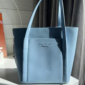 En ljusblå väska som är i läderimitation. Den är från MQs märke STOCKH LM STUDIO och det tillkommer ett axelband. Oanvänd!
