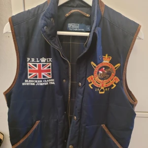 Väst Polo Ralph Lauren - Snygg mörkblå väst med bruna mockadetaljer. Grönrutig insida med innerfick. Hel och ren, snålt använd.