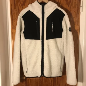 Sail racing patrol pile fleece - En vit teddyjacka från sail racing. Jacka har svarta detaljer och två svarta bröstfickor med dragkedja. Den är perfekt till vintern! I storlek L för män. Den är knappt använd men är ett år gammal.