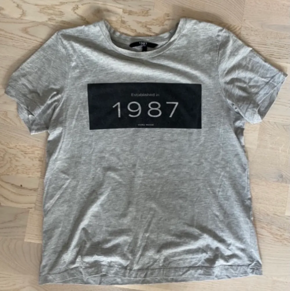 Säljer grå t-skirt från vero moda i strl M, passar S🌸 med tryck 1987 på💗 köpte för 299kr, säljer för 30kr💗 kan mötas i Sthlm eller fraktas🌸. T-shirts.