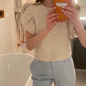 Söt topp - Söt topp med volanger på ärmarna från Zara. I ganska tjockt tyg och är nästan som sammets material. Väldigt skön, aldrig använd och lapparna är kvar! Beige/grå typ. Strl s men sitter mer som xs