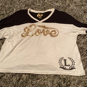 T-shirt med tryck strl 146-152 - Denna gulliga T-shirt har jag växt ur. Skönt material som kan användas som träningstopp eller vardagströja. Det står ’Love’ i guldiga paljetter på bysten samt ’L’ o ’Love’ längst ner i hörnet. Tröjan har inga fläckar eller skador. 