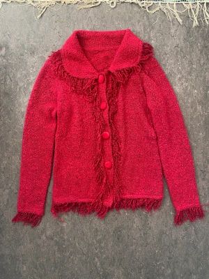 Röd kofta med fransar - Stickad röd kofta med fransar. Tyvärr för stor för mig. Nog bra på en M/L om den ska sitta tajt. Nedersta knappen saknas. Annars i gott skick. 