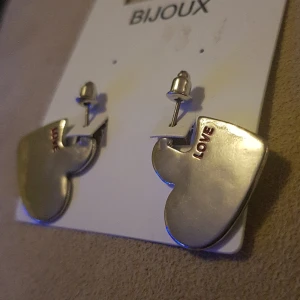 Hjärtformade örhängen  - Hjärtformade, silverfärgade örhängen med text LOVE. ALDRIG ANVÄNDA! 