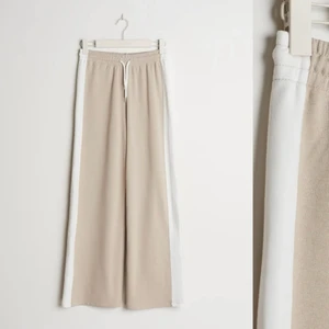 Sweatpants - Säljer dessa vida byxor i färgen beige med en vit rand på sidan från GinaTricot. Som har kommit till användning endast ett fåtal ggr. Går till anklarna på mig som är ungefär 165 cm.  Köpt för 349 kr. Frakten ingår ej i priset.  