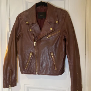 Vinröd jacka i lammskinn - Vinröd lammskinnsjacka med mässingsfärgade detaljer. Midjekort, supersnygg. Tyvärr skav på höger armbåge, men syns inte så mkt. Märket är Maje. 