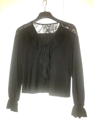 Svart blus med volang  - Volang och knytband vid urringningen, dekorativt spetsmönster högst upp i ryggen. Längd axelsöm/fåll 49cm