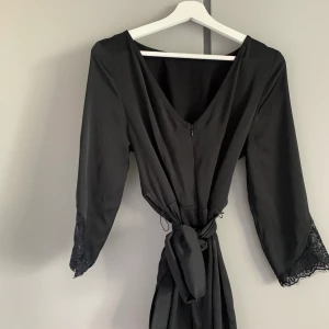 Svart klänning med spets och knyte - Klänning som har ett ”knyte i fram” som man knyter i bak av klänningen samt med spets i armarna. Storlek 36 som går precis över knäna på en person som är runt 170cm