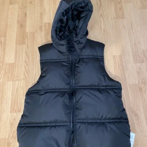 Svart puffetväst med luva - En oanvänd puffetväst från ASOS DESIGN. Bra skick och är som ny!
