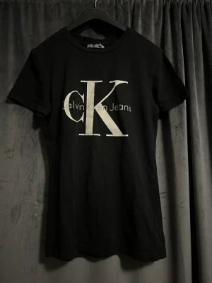 Calvin Klein T-shirt  - Svart Calvin Klein t-shirt med vit och grå logga. Använd flertal gånger, tyget är i bra skick dock har bokstäverna fått en aning med streck på vissa ställen som syns på bilderna. Storlek XS. Pris kan diskuteras. Köparen står för frakten. Betalning Swish.