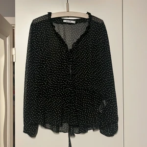 Blus från NA-KD - Svart Blus med vita prickar från NA-KD med knappar i mitten och band som går att knyta i midjan.  Storlek 38