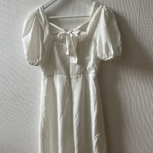 Vit klänning från Nelly x Glamorous - Supersöt vit knälång klänning med puffärmar, smockad detalj framtill och knytning i ryggen. Använd bara en gång. Passar perfekt till studenten bland annat:)