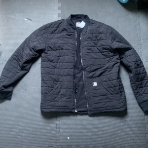 Svart quiltad bomberjacka Carhartt - Svart quiltad bomberjacka från Carhartt i storlek M. Jackan har en klassisk bomberkrage, dragkedja framtill och Carhartt-logga på framfickan Snygg och enkel design som funkar till det mesta.