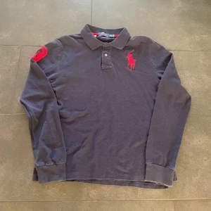 Mörkblå långärmad piké från Polo Ralph Lauren - Säljer en mörkblå långärmad pikétröja från Polo Ralph Lauren i slim fit. Tröjan har klassisk krage, två knappar, rött broderat polospelar-logo på bröstet och en röd siffra 3 på ärmen. Tillverkad i bomull, perfekt för en clean och sportig stil.