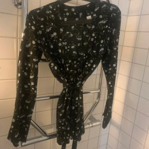 Svart blommig omlottkofta  - Supersöt svart kortkofta med vitt blommönster och långa ärmar. Omlottmodell med knyt i midjan som ger en snygg siluett. Perfekt för dig som gillar en feminin och trendig stil.
