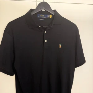 Svart pikétröja från Polo Ralph Lauren - Klassisk svart pikétröja från Polo Ralph Lauren med broderad logga på bröstet. Tröjan har korta ärmar, krage och två knappar framtill. Perfekt för en clean och stilren look.