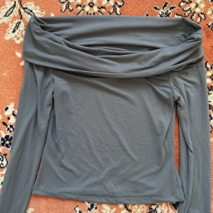Grå offshoulder topp med lång ärm - Snygg grå offshoulder topp med långa ärmar och bred vikt kant över axlarna. Toppens passform är tight och den är lätt croppad, perfekt till jeans eller kjol. Mjuk och stretchig i materialet för en bekväm känsla.