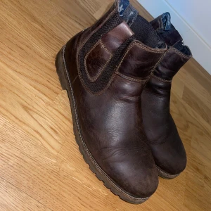 Bruna chelsea boots från U.S. Polo Assn. - Snygga bruna chelsea boots från U.S. Polo Assn. i äkta skinn. Klassisk rund tå, resår på sidorna och robust sula. Perfekta för höst och vinter, med diskreta logodetaljer på insidan och dragflikar upptill för enkel påtagning.