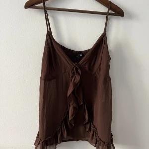 Brun vintage volangtop från H&M - Säljer en brun linnetopp från H&M med tunna axelband och v-ringning. Toppen har volangdetaljer framtill och längs nederkanten samt en snygg knytning vid bysten. Lätt och luftig känsla, perfekt för varma dagar.