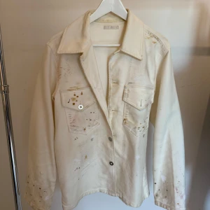 Vit overshirt från Our Legacy - Säljer en vit overshirt från Our Legacy i 100% bomull med handgjorda färgstänk och unika detaljer. Jackan har två stora bröstfickor med lock och knappar, klassisk krage och lång ärm. Perfekt för dig som gillar en kreativ och individuell stil.