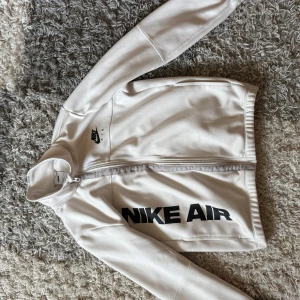 Vit Nike Air hoddie med dragkedja - Snygg vit Nike Air hoddie i storlek 147 -158 cm  med hel dragkedja framtill och svart logga på bröstet samt stort tryck längs sidan. Hoodie har hög krage och är tillverkad i ett mjukt, lätt material som passar perfekt till streetwear eller träning.