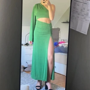 Grön långklänning med slits och oneshoulder - Snygg grön långklänning med en ärm och oneshoulder-design. Klänningen har en hög slits på ena sidan och en tight passform upptill med rynkade detaljer vid höften. Perfekt för dig som vill sticka ut med en unik och trendig look.
