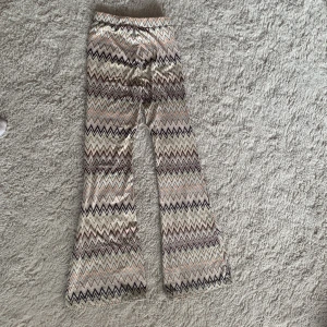 Mönstrade bootcut byxor från KappAhl - Säljer ett par riktigt snygga bootcut byxor från KappAhl med zigzag-mönster i beige, svart, brun och ljusrosa toner. Byxorna har hög midja och är gjorda i ett mjukt, stretchigt material som sitter skönt på kroppen. Perfekta för dig som vill sticka ut lite extra.