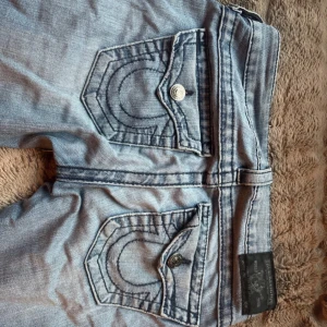 Ljusblå bootcut jeans från True Religion - Säljer ett par ljusblå jeans från True Religion med bootcut passform. Jeansen har snygga kontrastsömmar, stora bakfickor med lock och knappar samt klassisk logga. Tillverkade i jeansmaterial med en cool tvättad look. innerbenslängd:80cm midjemått:35cm