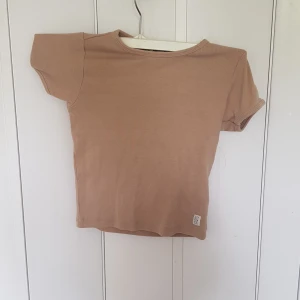Beige ribbad topp från Ett Fem Sju - Snygg beige ribbad topp från Ett Fem Sju med korta ärmar och rund halsringning. Enkel och stilren design som passar perfekt till jeans eller kjol. Mjuk och stretchig bomullskvalitet för en skön känsla hela dagen.