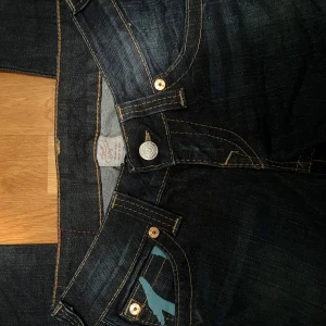 True Religion jeans med blå detaljer - Mörkblå jeans från True Religion med ikoniska blå broderade detaljer på bakfickorna och kontrastsömmar. Klassisk femficksmodell med raka ben och silverfärgade knappar. 