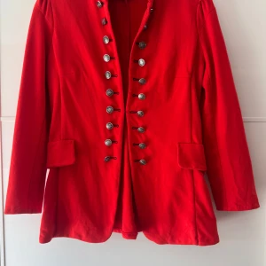 Röd rock med dekorativa knappar - Snygg kavaj med långa ärmar, som jag kortade till 54,5cm och dubbla rader av silvriga knappar framtill. Jackan har en rak, lite längre passform. Perfekt för dig som vill sticka ut med en statement-jacka. Materialet känns mjukt och har en klassisk look. Storlek L men är förminskad som M, bröst 94cm, längd 68cm. Lite nopprig under ärmarna men det syns inte.