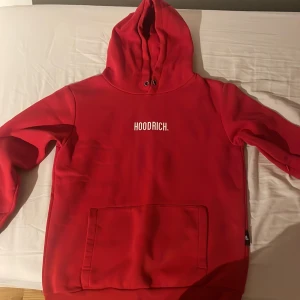 Röd hoodie från Hoodrich med tryck - Röd hoodie från Hoodrich med vit logga på bröstet. Klassisk känguruficka framtill, huva med snörning och långa ärmar. Perfekt för dig som gillar streetwear och vill sticka ut med en färgstark look.
