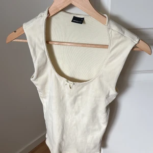 Ljusbeige ribbad linnetopp Gina Tricot - Supersöt ljusbeige linnetopp från Gina Tricot med ribbad struktur och liten rosett framtill vid urringningen. Toppen är ärmlös och har en klassisk, enkel design som är lätt att matcha med allt. Perfekt för varma dagar eller lager på lager.
