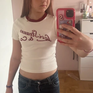 Vit Levi's t-shirt med röd text - Vit croppad t-shirt från Levi's med röd text 'Levi Strauss & Co.' framtill och matchande röd kant vid halsen. T-shirten har korta ärmar och är i mjuk bomull, perfekt till jeans eller shorts för en chill look. Är dessutom aldrig använd.