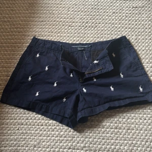 Mörkblå lågmidjade shorts från Ralph Lauren - Snygga lågmidjade mörkblå shorts från Ralph Lauren med broderade vita polospelare över hela tyget. Klassisk passform med dragkedja och knäppning, samt fickor både fram och bak. Perfekta för sommardagar och chill häng.