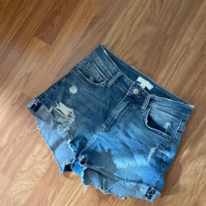 Blå slitna jeansshorts med fransar - Snygga blå jeansshorts med hög midja och råa, fransiga kanter. Shortsen har slitningar och hål framtill för en cool, avslappnad look. Klassisk femficksmodell med bälteshällor och knappstängning. Perfekta för sommardagar och festivaler.