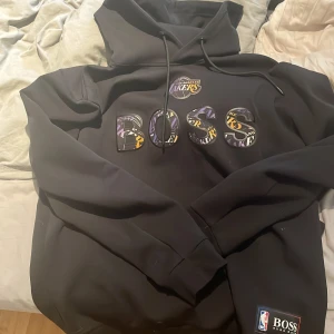 Svart Lakers hoodie från Hugo Boss - Snygg svart hoodie från Hugo Boss x NBA med Los Angeles. storleken är XL men jag själv har M/L och den sitter bra på mig som är 185cm, 79kg.